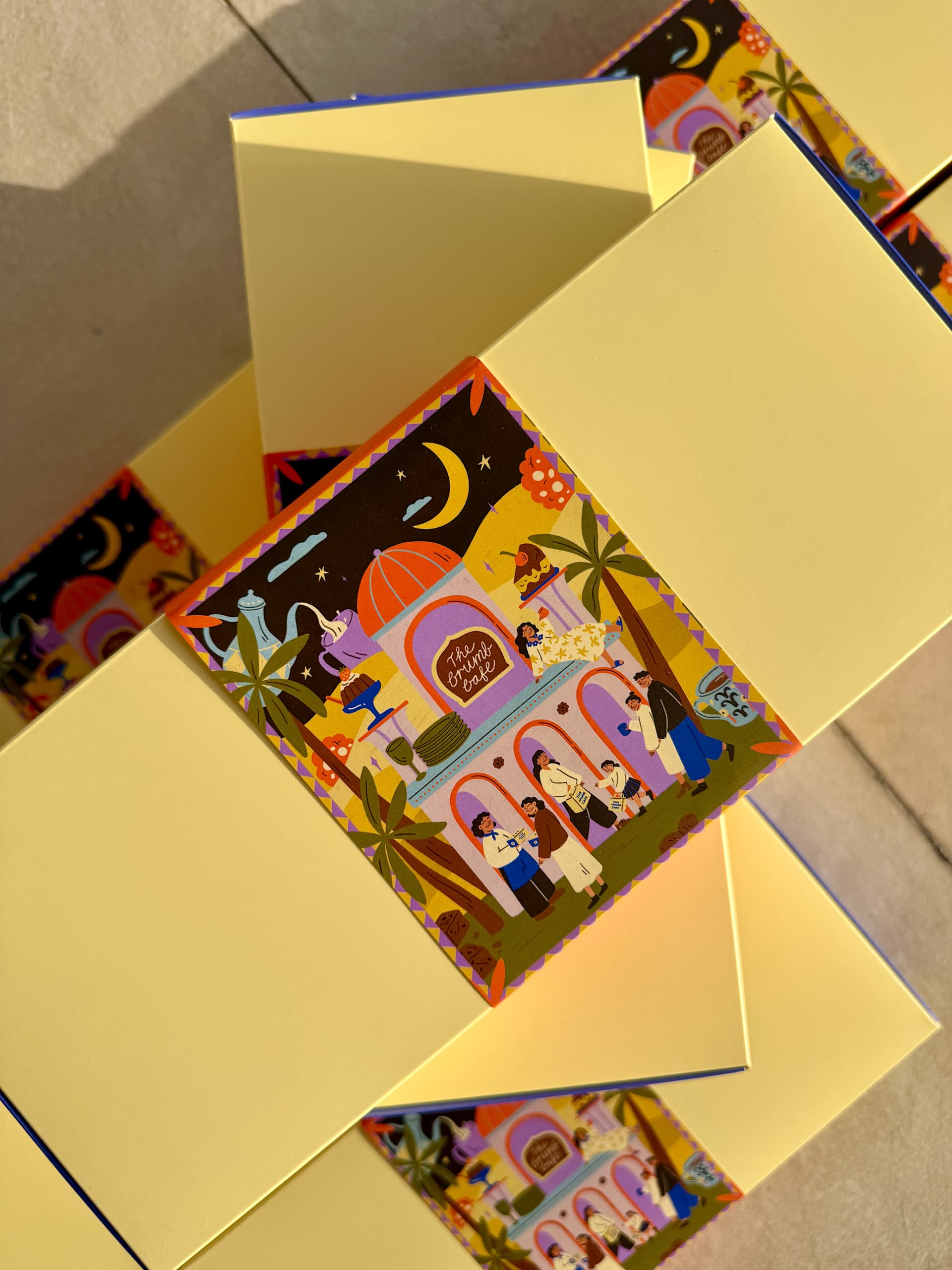 Cream-colored packaging boxes wrapped with illustrated Ramadan sleeve packaging featuring crescent moons, palm trees and a vibrant market scene with people gathered around a domed building.