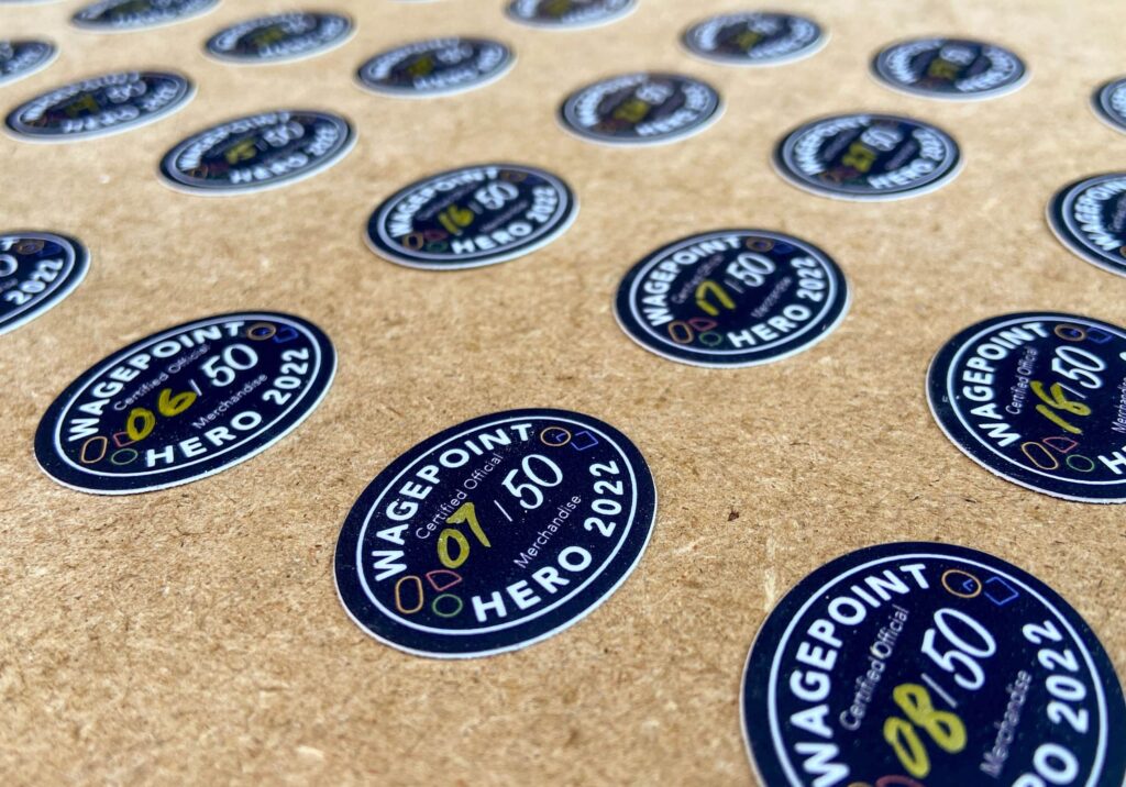 Rows of custom small, round, die-cut sticker singles with a frame, the &ldquo;Wagepoint hero 2022&rdquo; text inside the frame and golden hand-written numbers &ldquo;&ndash;/50&rdquo;]