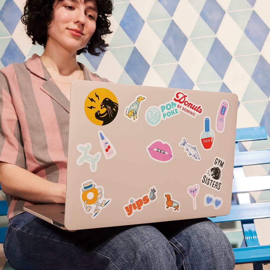 A woman sitting in front of a laptop decorated with a variety of branded stickers, micro-merch from different brands: Pon Poke, Yips, Gym Sister, Donuts by Dominic, Dew and more