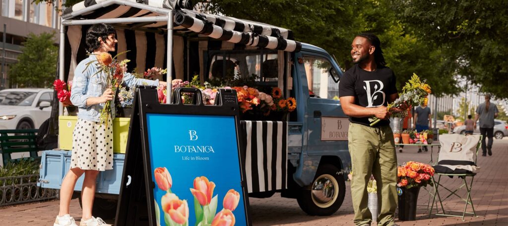 An A-frame sign featuring flowers, brand logo and tagline “Life in bloom” standing in front of a popup van.