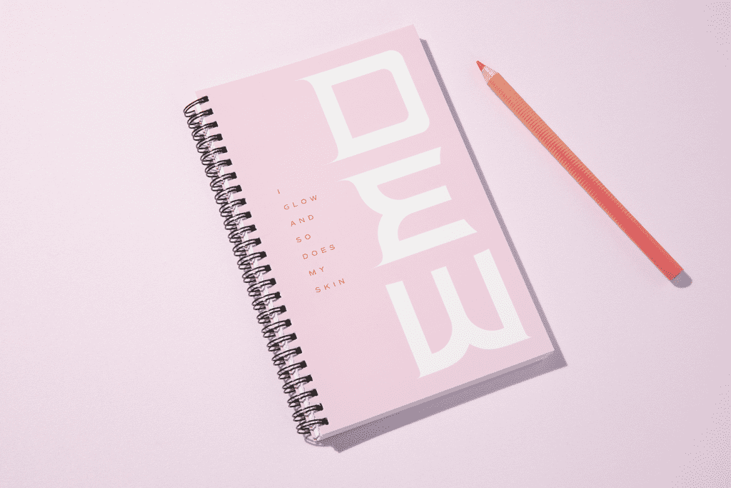 Pink spiral-bound notebook with white typography next to a red-orange pencil