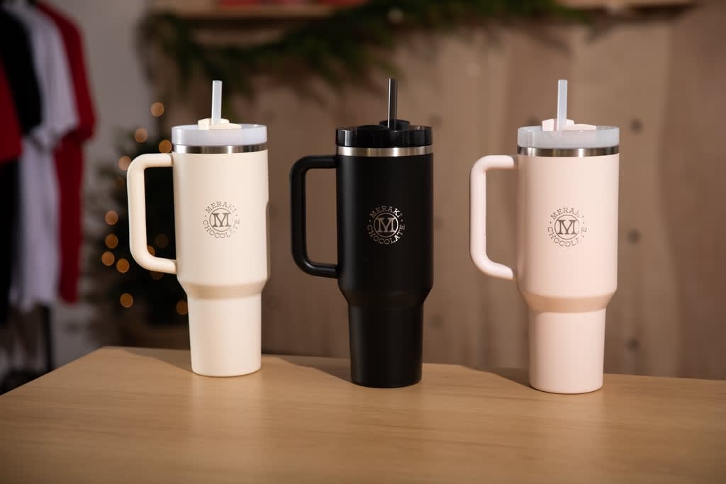 Three branded tumblers in cream, black and baby pink with small logo.