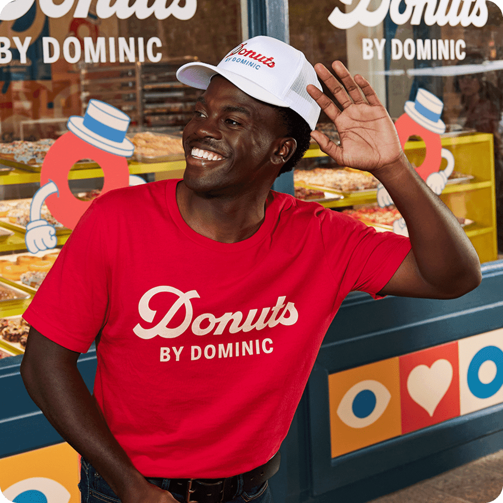 A donut shop employee wearing a branded uniform: an embroidered cap and a red T-shirt with a full chest logo design placement
