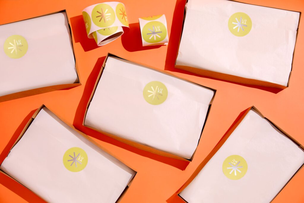 Open cardboard packaging boxes with products wrapped in white tissue paper and sealed with round LUXA logo stickers, arranged on an orange background.