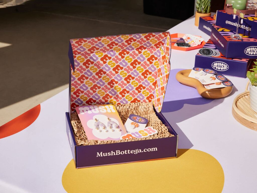 Open purple custom jewelry packaging box with colorful patterned interior, crinkle paper filler, earrings, branded cards and stickers from Mush Bottega.