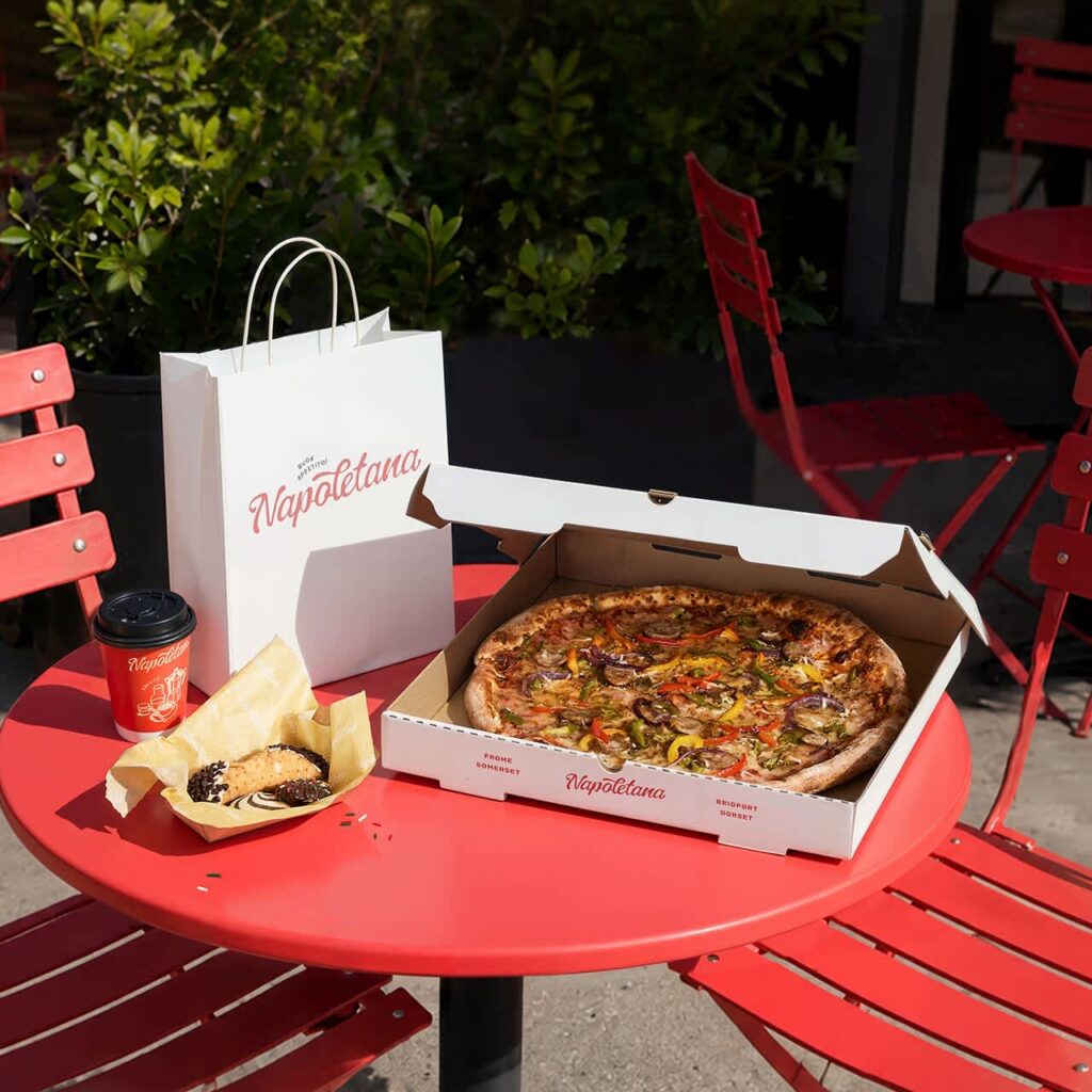 Open custom pizza box with a freshly baked pizza on a red outdoor café table, alongside branded takeaway packaging and food.
