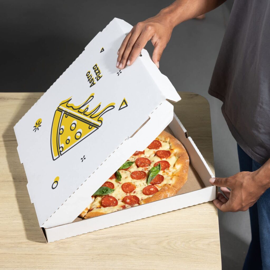 Person opening a white custom pizza box with a yellow pizza slice illustration, revealing a pepperoni pizza topped with fresh basil.