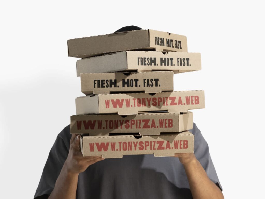 Person holding a tall stack of kraft pizza boxes with bold slogans and the restaurant's website printed along the side rims.