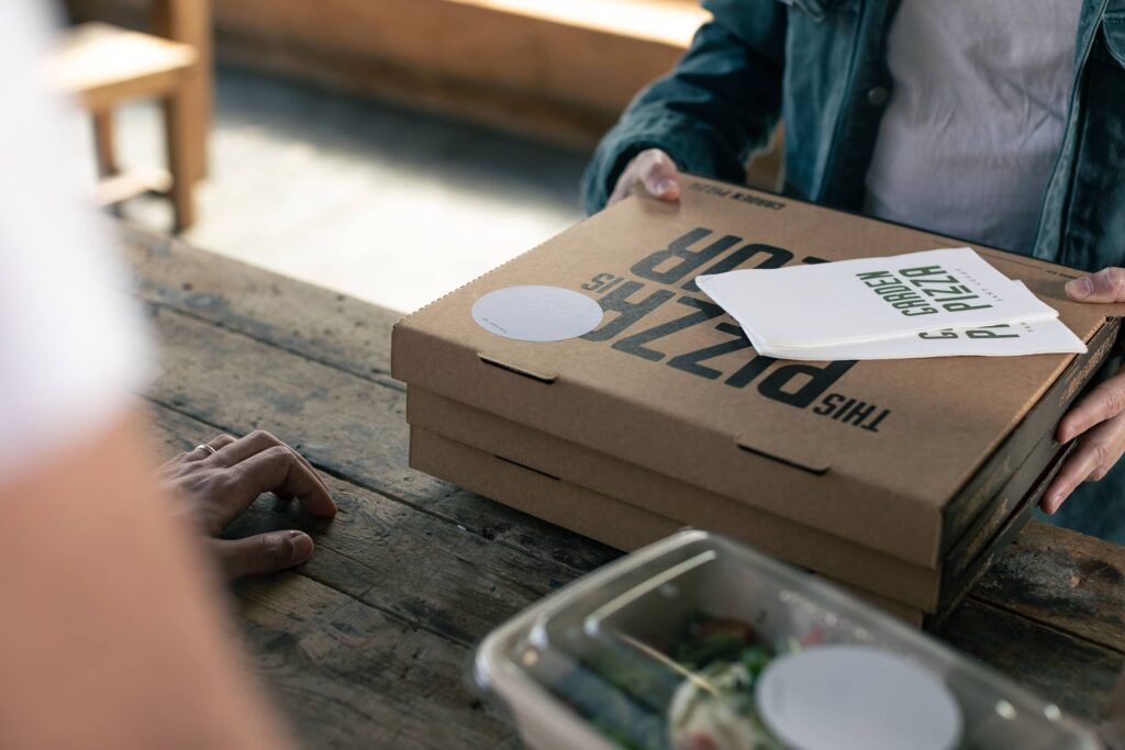 Two stacked kraft pizza boxes with ventilation holes along the sides, featuring bold black branding and branded napkins, being handed to a customer.
