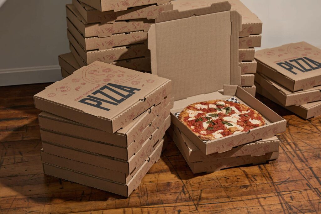 Stacks of custom kraft pizza boxes on a wooden table, with one open box revealing a freshly baked pizza topped with mozzarella, tomato and basil.