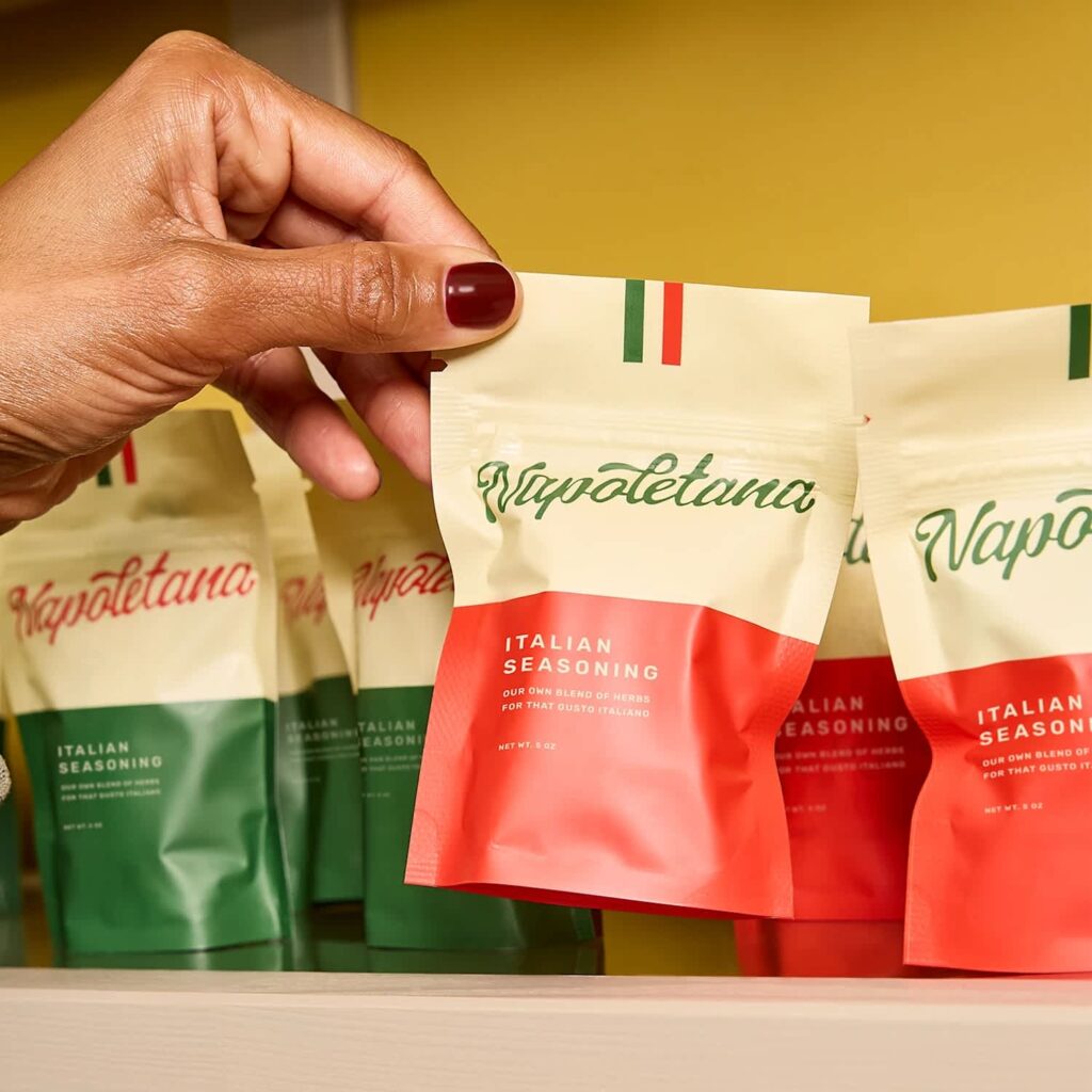 Hand holding a red and cream Vapolettano Italian seasoning stand-up pouch, with matching green and red custom pouches displayed behind it.