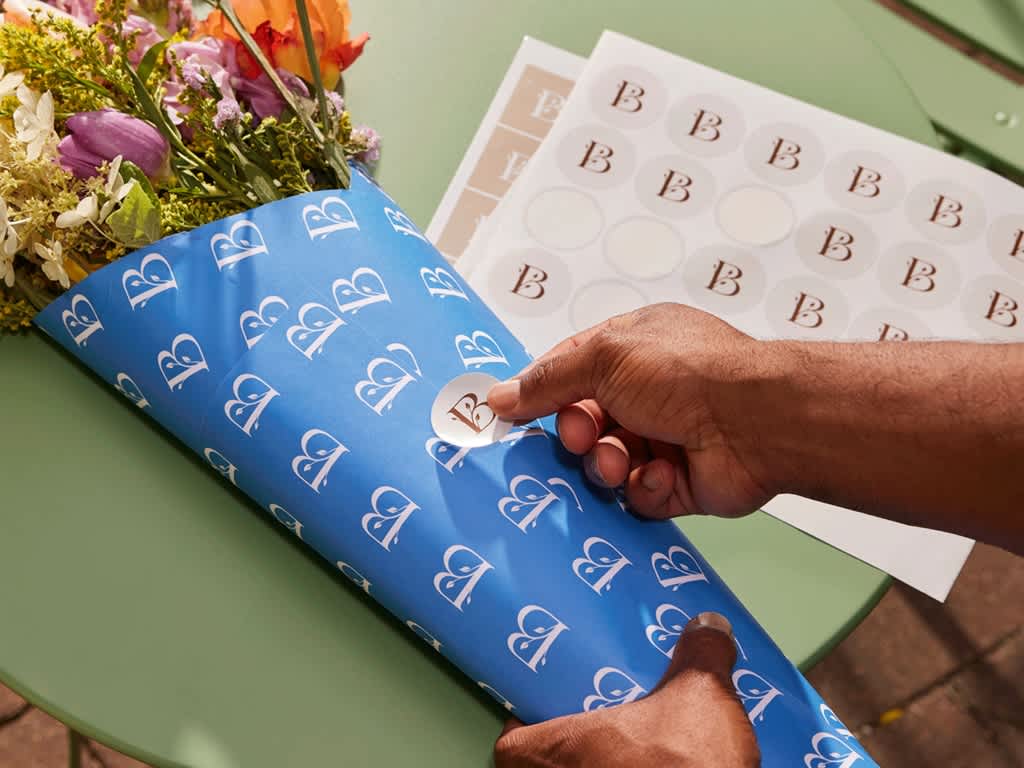 Person sealing blue branded tissue paper with a round logo sticker, with sheets of matching custom stickers and flowers in the background.