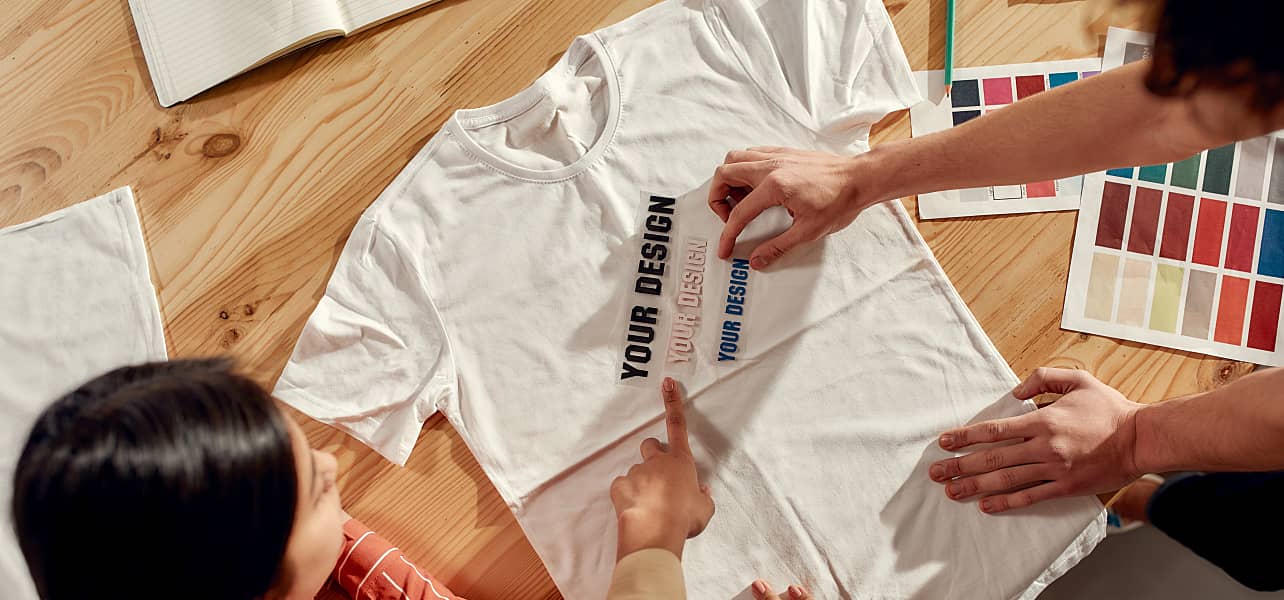 Top view of creative people trying on stickers with text, while discussing logo and design of T-shirt. Young man and women working together at custom T-shirt, clothing printing company. Cropped shot