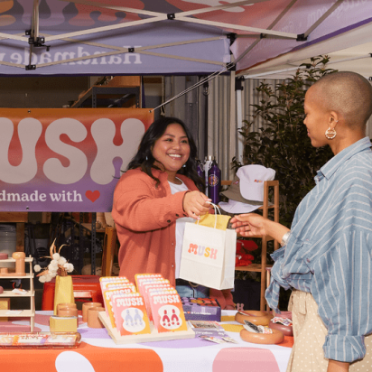 A vibrant craft fair stand where a smiling vendor hands a branded shopping bag to a happy customer. A real-world example of how to do market research for a small business.