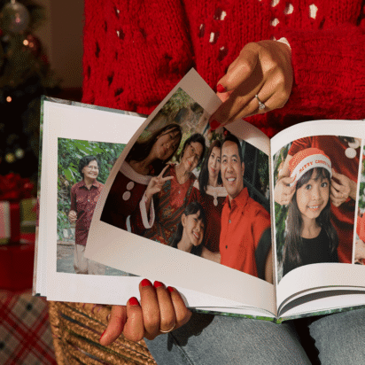 Woman holding a photo book with family Christmas pictures, surrounded by presents and a Christmas tree — personalized Christmas gifts idea.
