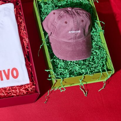 Three products in festive packaging, with the visible branding "Apertivo", are laid out side by side on a red table: A beanie, a thsirt and a cap