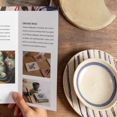 Hands holding a tri-fold brochure on ceramic workshops, surrounded by handmade pottery on a wooden table – example of brochure folds in real use.
