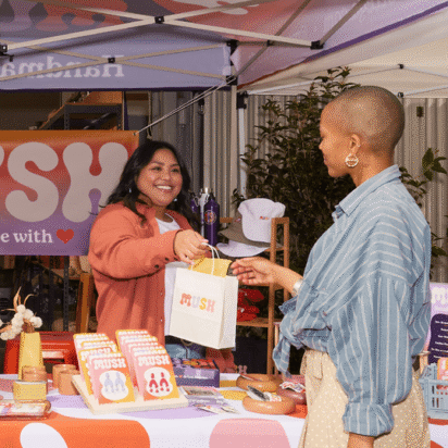 Small business owner handing a branded bag to a customer at a colorful outdoor market booth during a pop-up event