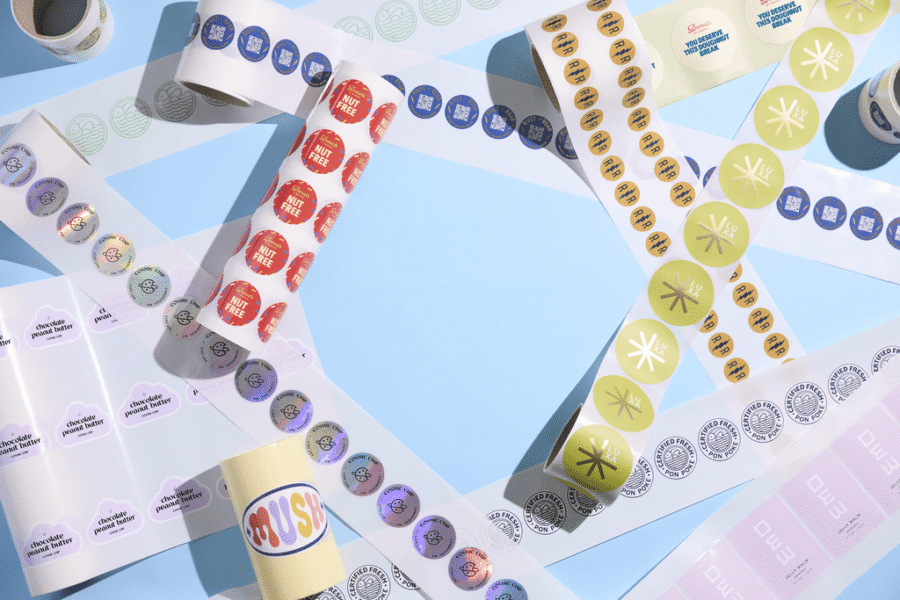 Various custom logo stickers on rolls in multiple styles, colors and finishes arranged on a pastel blue surface