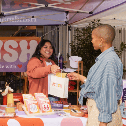 Small business owner handing a branded bag to a customer at a colorful outdoor market booth during a pop-up event