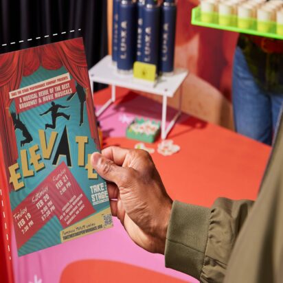 Person holding a colorful event flyer with bold typography and performance details at a promotional table