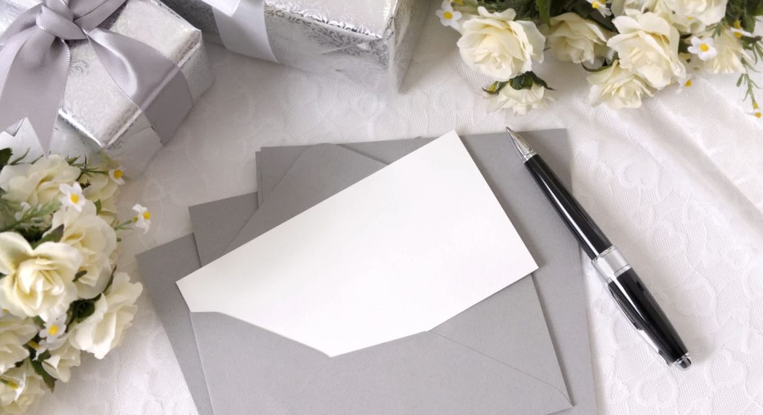 Writing paper or wedding invitation with envelope laid on bridal lace with several wedding gifts and white rose bouquets. Space for copy.