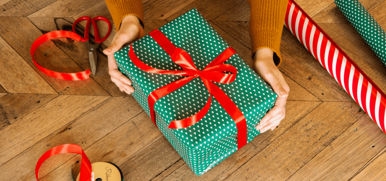 Two hands holding a green wrapped gift with a red bow on a wooden surface.