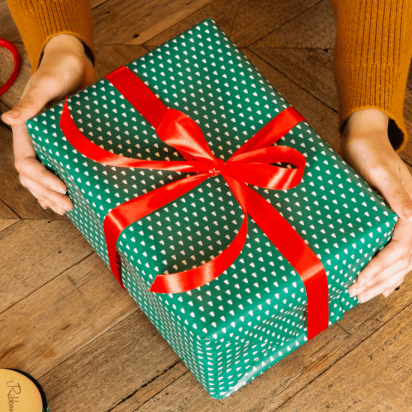 Two hands holding a green wrapped gift with a red bow on a wooden surface.