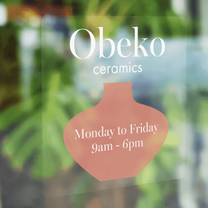 Storefront with wooden decor and indoor plants in the background, featuring a transparent logo sticker with business hours on the glass.