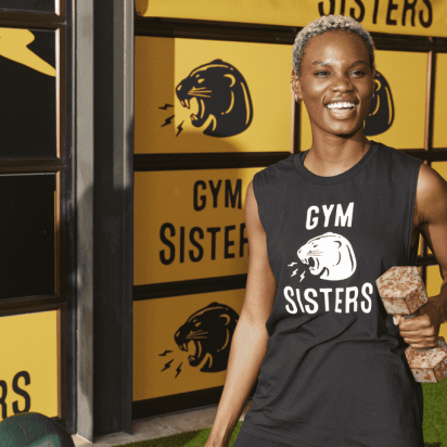 Gym woman lifting a weight while wearing a branded Gym Sisters T-shirt, with a gym wall decorated with stickers and a banner in the background.