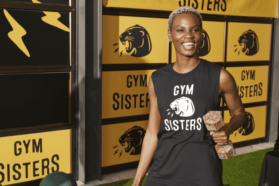 Gym woman lifting a weight while wearing a branded Gym Sisters T-shirt, with a gym wall decorated with stickers and a banner in the background.