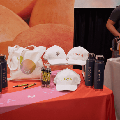 Colorful trade show table displaying branded swag items like hats, bottles, tote bags, and pens with a company logo.