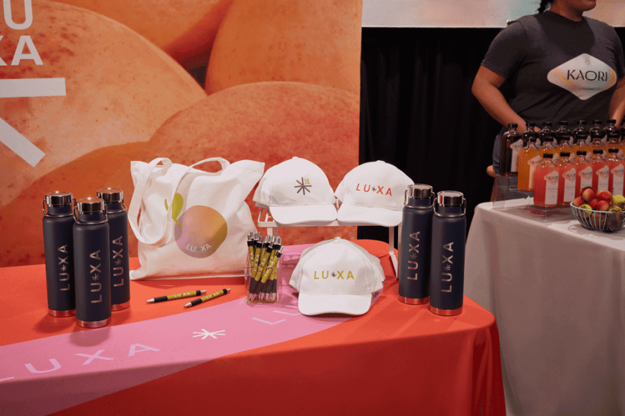 Colorful trade show table displaying branded swag items like hats, bottles, tote bags, and pens with a company logo.