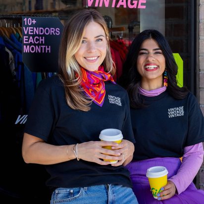 Two women wearing branded T-shirts and holding takeout drink cups in front of their store window.