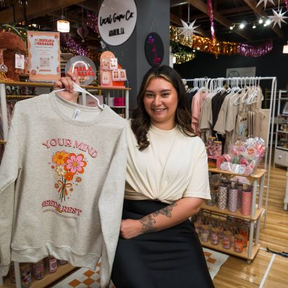 Smiling business owner showing a custom t-shirt inside her clothing and accessories store — signs of a successful business.