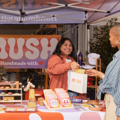 A vibrant craft fair stand where a smiling vendor hands a branded shopping bag to a happy customer. A real-world example of how to do market research for a small business.