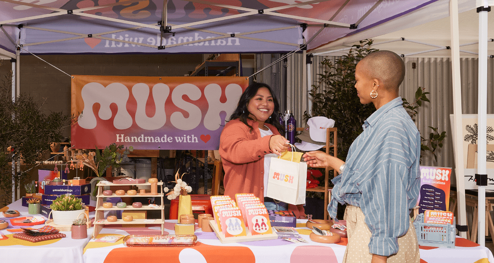 A vibrant craft fair stand where a smiling vendor hands a branded shopping bag to a happy customer. A real-world example of how to do market research for a small business.