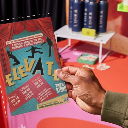 Person holding a colorful event flyer with bold typography and performance details at a promotional table