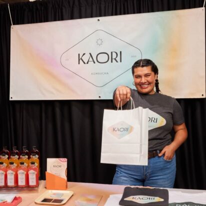 Smiling small business owner at trade show stand holding branded paper bag, with neatly displayed products and flyers.