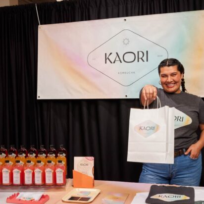 Small business owner at a fair stand holding a custom paper bag with her logo, colorful products on the table and a branded community banner in the background.