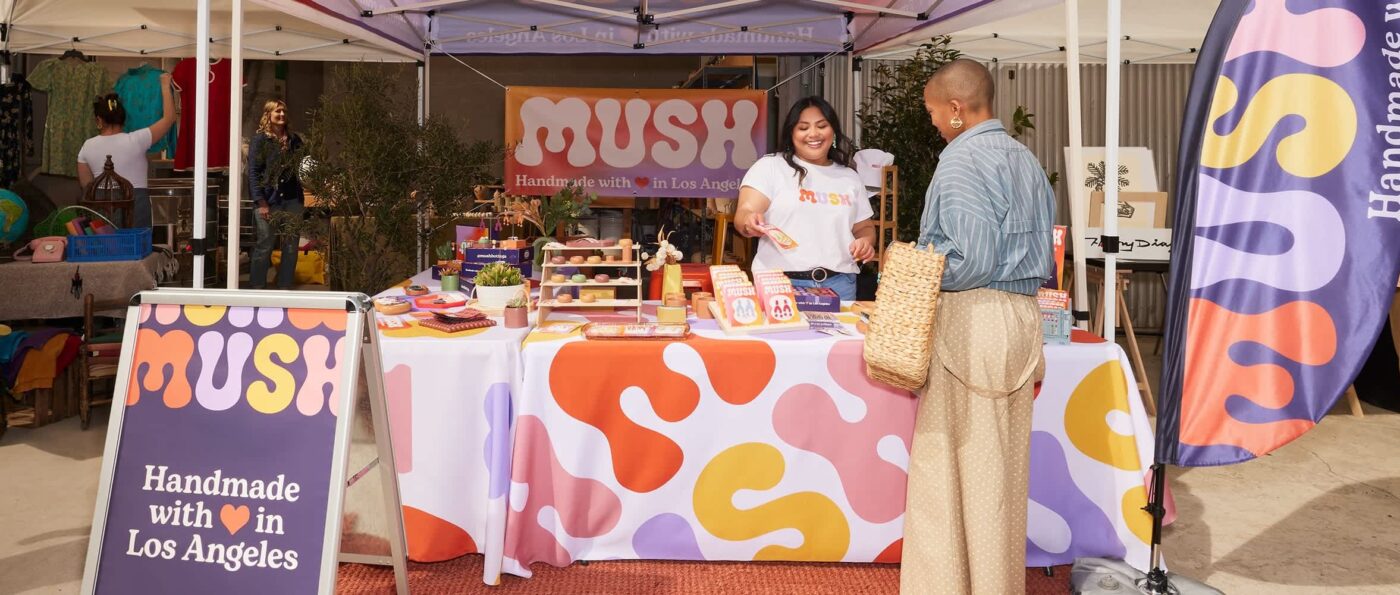 Outdoor fair stand showcasing signage in retail stores, with coordinated banner, vertical flag, A-sign and smiling vendor and customer.