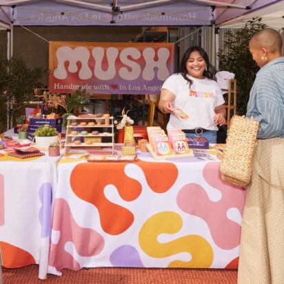 Outdoor fair stand showcasing signage in retail stores, with coordinated banner, vertical flag, A-sign and smiling vendor and customer.