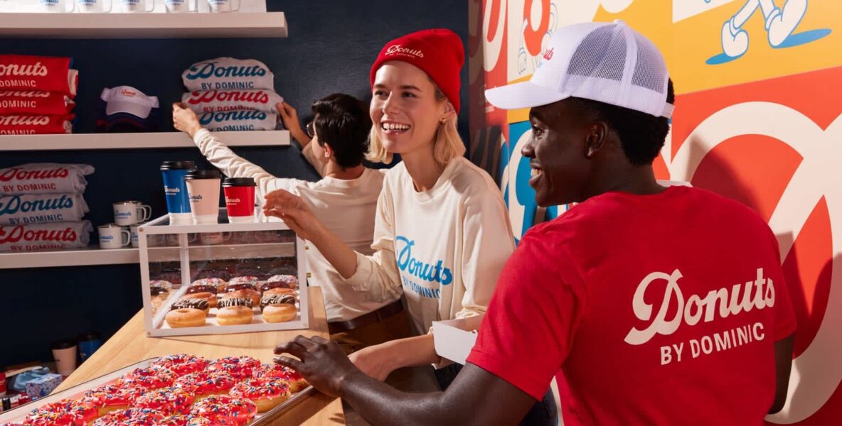 Two smiling employees behind the counter of a colorful donut shop, wearing branded accessories – part of the VistaPrint Small Business Happiness Report.
