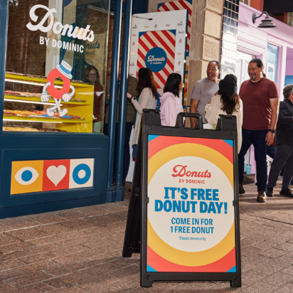 Colorful A-frame sign outside a donut shop with people lined up to enter.