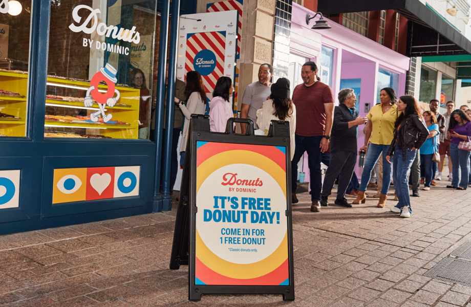 Colorful A-frame sign outside a donut shop with people lined up to enter.