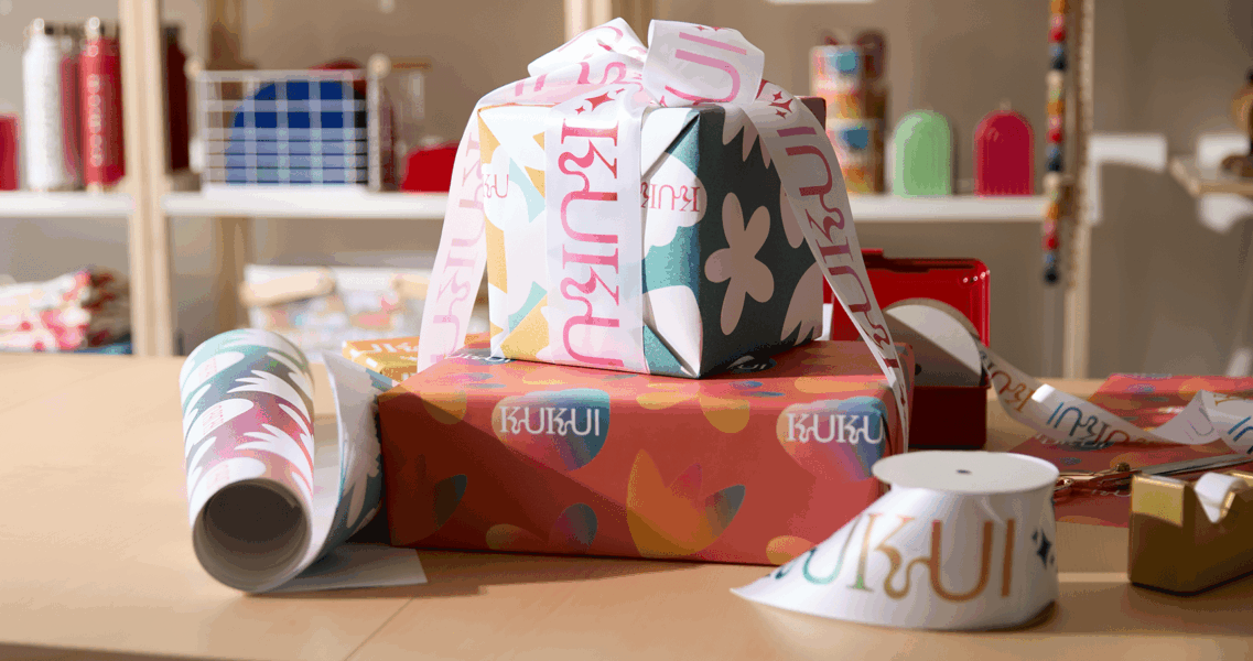 Holiday giveaway preparation with branded wrapping paper and ribbon on a desk, featuring tape and packaging materials.