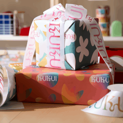 Holiday giveaway preparation with branded wrapping paper and ribbon on a desk, featuring tape and packaging materials.