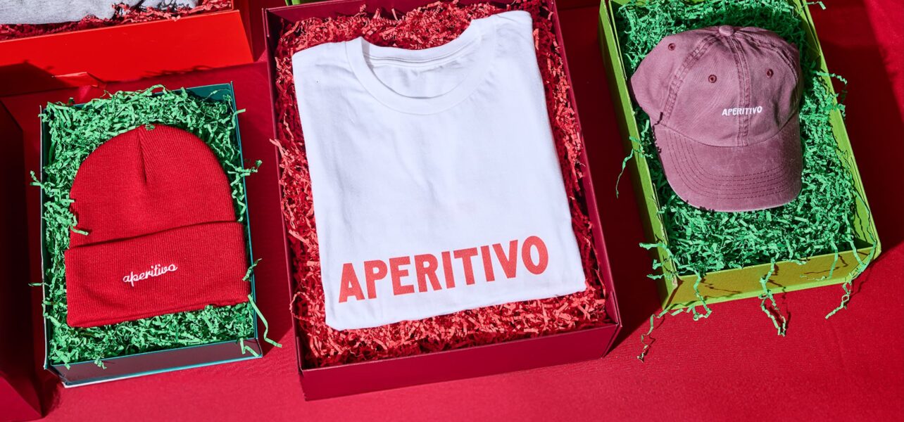 Three products in festive packaging, with the visible branding "Apertivo", are laid out side by side on a red table: A beanie, a thsirt and a cap