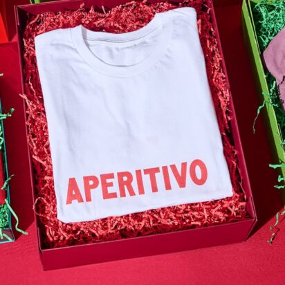 Three products in festive packaging, with the visible branding "Apertivo", are laid out side by side on a red table: A beanie, a thsirt and a cap