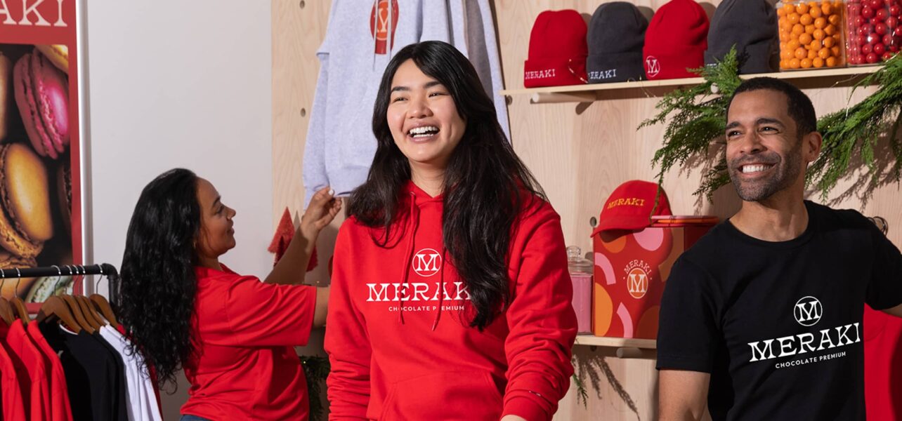 Smiling staff wear red coloured merch for Meraki (chocolate premium) as they stock a store's shelves.